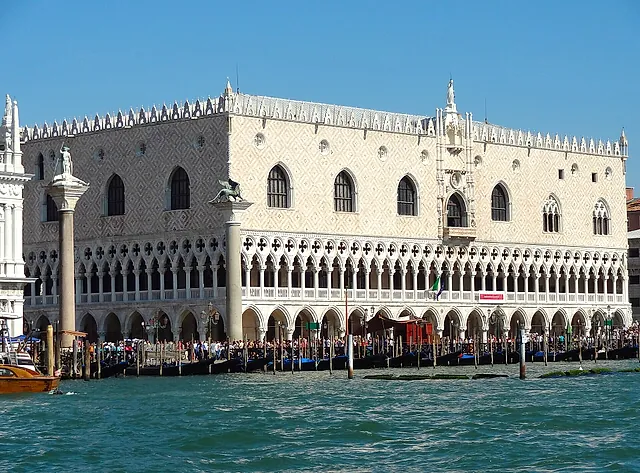 La fachada gótica del Palacio Ducal junto al muelle, con arcadas blancas y un muro de dibujos rosas y blancos bajo un cielo azul